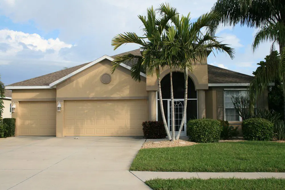 A modern, single-story house with a light beige exterior, a three-car garage, and a well-maintained lawn. The driveway is flawlessly paved by Reno Concrete Solutions. There are two tall palm trees at the front and a covered entryway with a screened door. The sky in the background is partly cloudy.