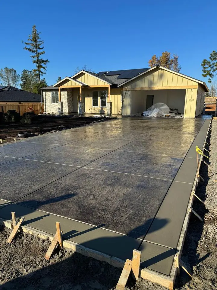 A long, freshly wet concrete driveway bordered by a low red brick wall and green bushes leads to a beige two-story house. Several large items covered with tarps line the right side of the driveway. Yellow caution tape is visible in the foreground, hinting at ongoing work by Reno Concrete Solutions.