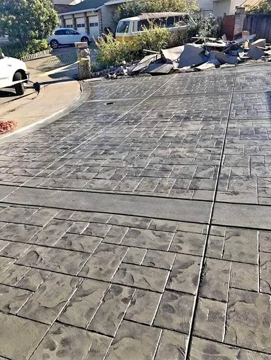 A large, modern single-story house with a stone exterior and dark wooden garage doors. The house features a well-manicured lawn, a spacious driveway with a decorative border by Reno Concrete Solutions, and a blue sky with scattered white clouds above. Contact us for your free service quote today!