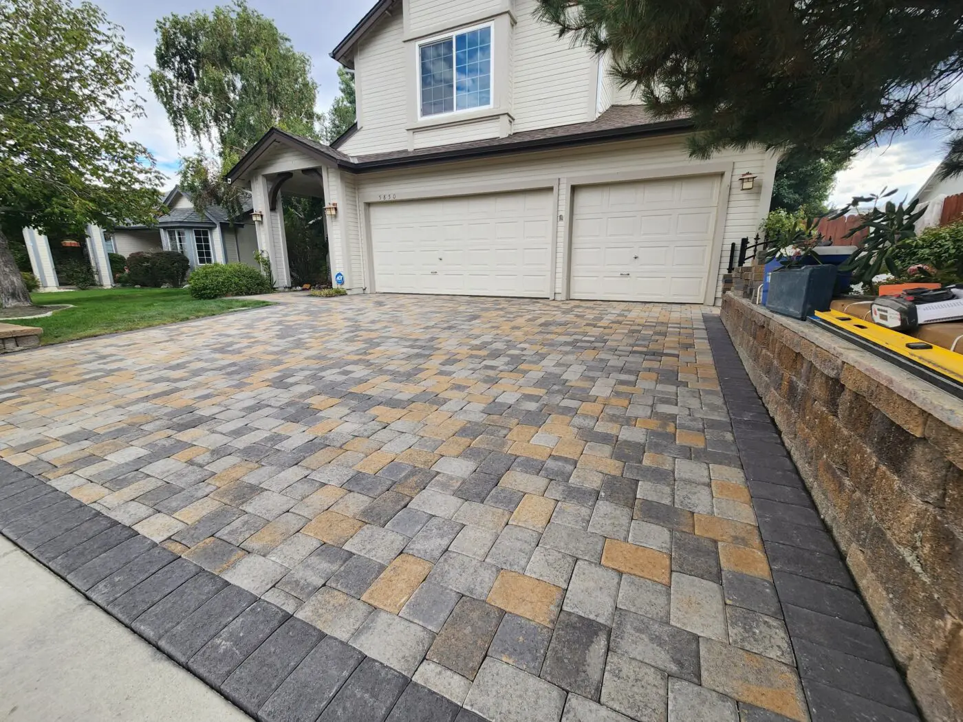 A backyard features a stone-paved pathway, showcasing expert pavers installation, leading to a lawn with desert plants and trees. To the right is part of a gray surface, possibly a pool or court. The landscape is under a clear blue sky with scattered clouds.